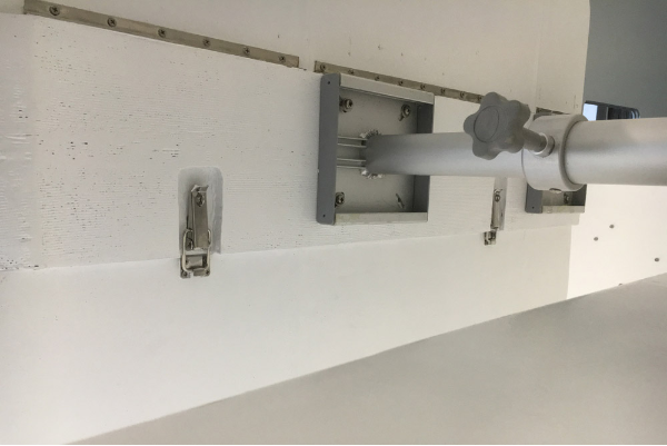 Underneath. Hinge on one side, two tensor latches on the other